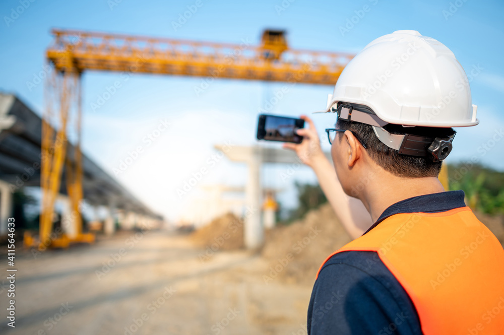 Smart Asian worker man or male civil engineer with protective safety helmet and reflective vest using using smartphone for taking photo at construction site. Smart Asian worker man or male civil engineer with protective safety helmet and reflective vest using using smartphone for taking photo at construction site.