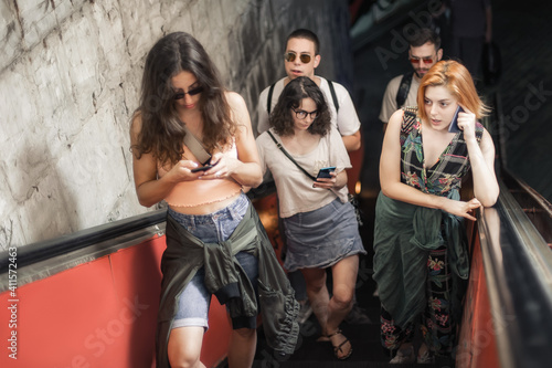 Group of friends talk on smartphones while standing on escalator. Modern people with cell phones outdoor. Mobile communication technology. High angle view