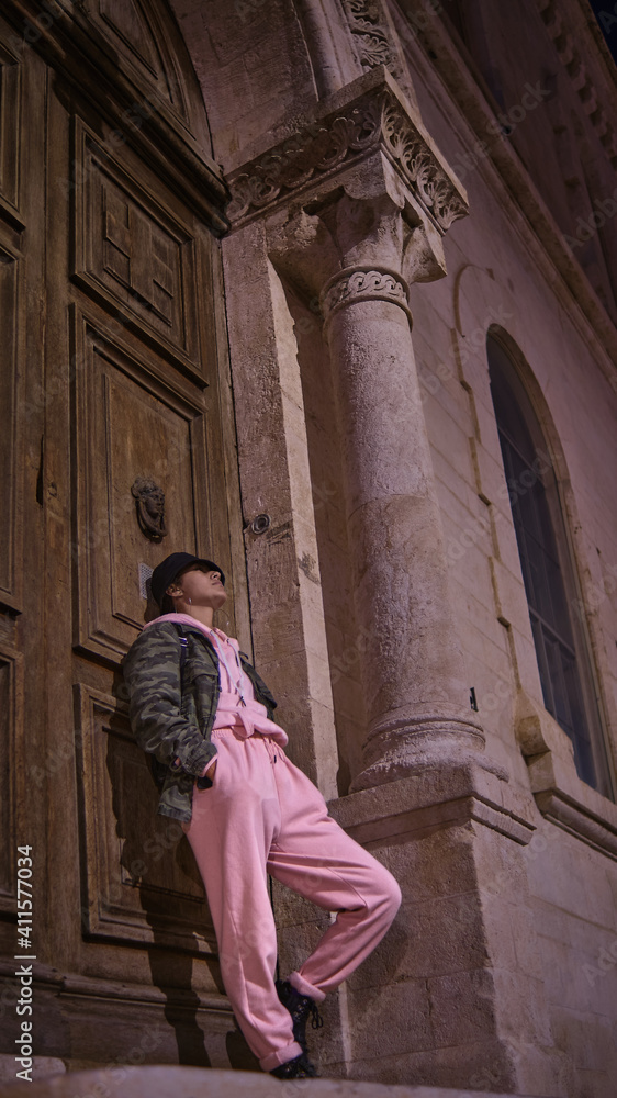 latina woman with black hat leaning against a large wooden door of an old building, wearing pink sport pants and camouflage jacket