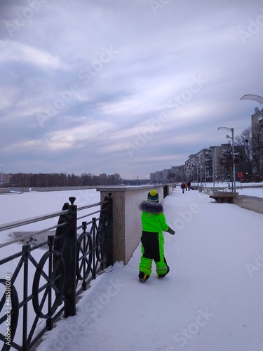 Boy in green ski overalls on the embankment of the river in winter on a snowy day
