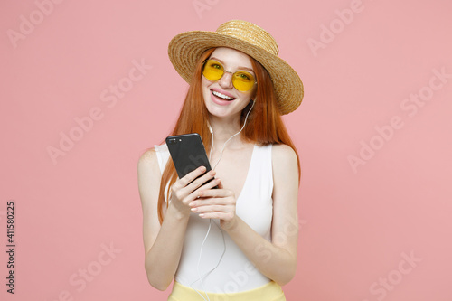 Young smiling redhead ginger student woman 20s wearing straw hat glasses summer clothes holding mobile cell phone listening to music with headphones isolated on pastel pink background studio portrait.