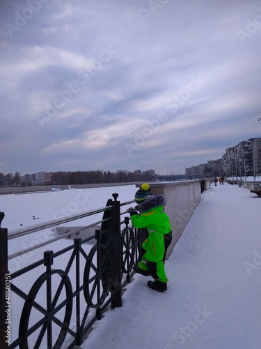 Boy in green ski overalls on the embankment of the river in winter on a snowy day