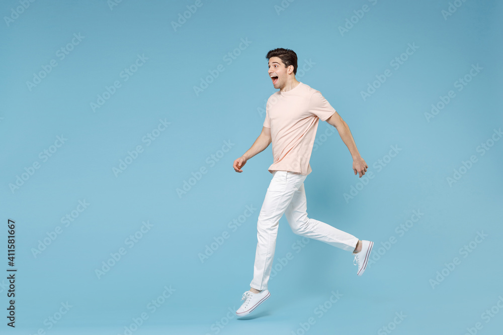 Full length side view of young caucasian attractive surprised happy excited student happy man 20s wear beige t-shirt, white pants sneackers running isolated on blue color background studio portrait.