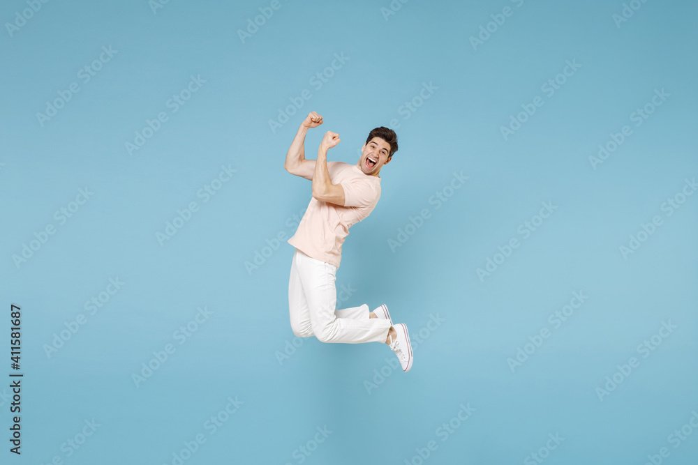 Full length of young caucasian happy excited overjoyed student man 20s in beige t-shirt white pants do winner gesture clench fists in air jump high scream isolated on blue background studio portrait. Full length of young caucasian happy excited overjoyed student man 20s in beige t-shirt white pants do winner gesture clench fists in air jump high scream isolated on blue background studio portrait.