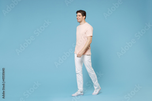 Full length of young caucasian attractive fashionable smiling student man 20s wearing beige t-shirt white pants sneackers walking going looking aside isolated on blue color background studio portrait © ViDi Studio
