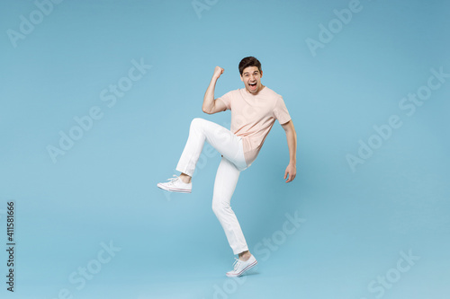 Full length of young caucasian happy excited successful student man in beige t-shirt white pants do winner gesture clench fists  raise up leg look camera isolated on blue background studio portrait.