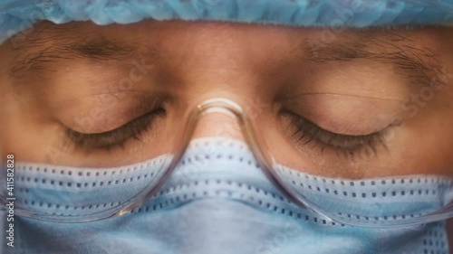 Hospital worker male in medical suit and protective glasses close up. Sad and tired medic opens eyes and looking atraight at camera.