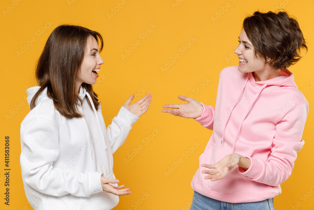 Excited funny two young women friends 20s wearing casual white pink hoodies standing speaking talking spreading hands looking at each other isolated on bright yellow color background studio portrait. Excited funny two young women friends 20s wearing casual white pink hoodies standing speaking talking spreading hands looking at each other isolated on bright yellow color background studio portrait.