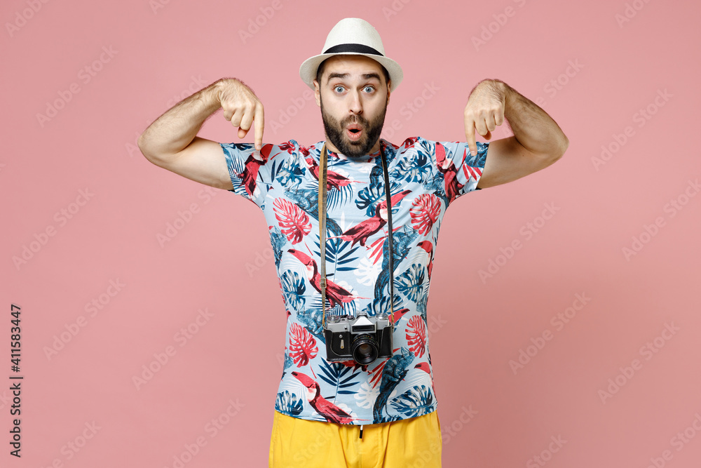 Shocked young traveler tourist man in summer clothes hat with photo camera pointing index fingers down isolated on pink background. Passenger traveling abroad on weekends. Air flight journey concept.