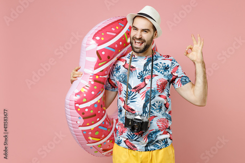 Laughing young traveler tourist man in summer clothes hat photo camera hold inflatable ring showing ok gesture isolated on pink background. Passenger traveling on weekends. Air flight journey concept.