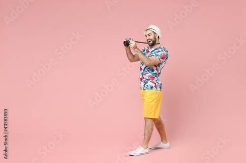 Full length side view of funny traveler tourist man in summer clothes hat taking pictures on vintage photo camera isolated on pink background. Passenger travel on weekends. Air flight journey concept.