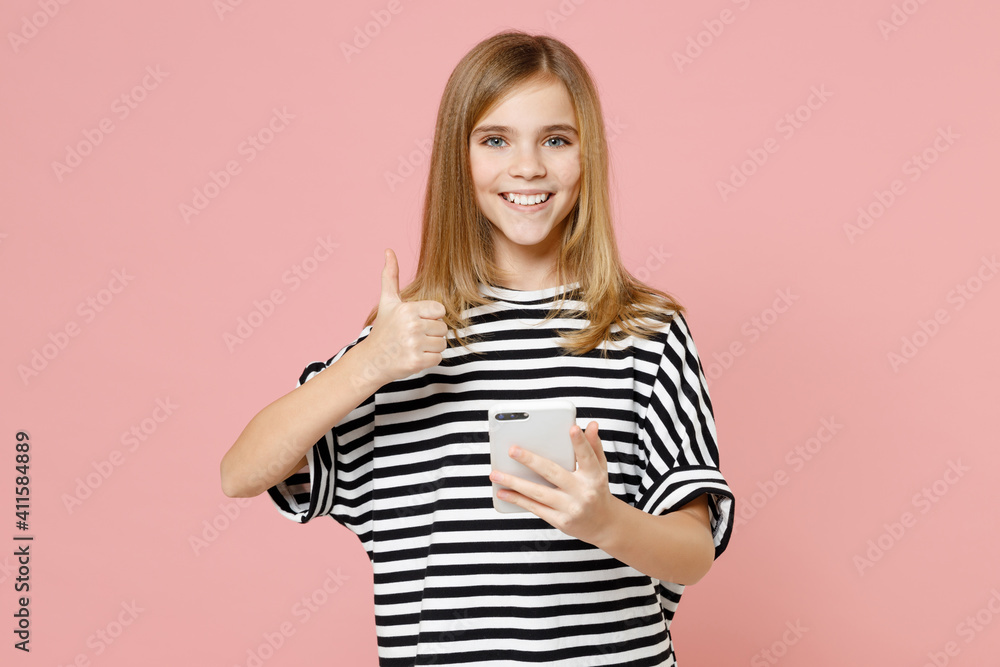 Little blonde happy kid girl 12-13 years old in striped oversized t-shirt hold mobile cell phone chat show thumb up gesture isolated on pastel pink background children. Childhood lifestyle concept. Little blonde happy kid girl 12-13 years old in striped oversized t-shirt hold mobile cell phone chat show thumb up gesture isolated on pastel pink background children. Childhood lifestyle concept.