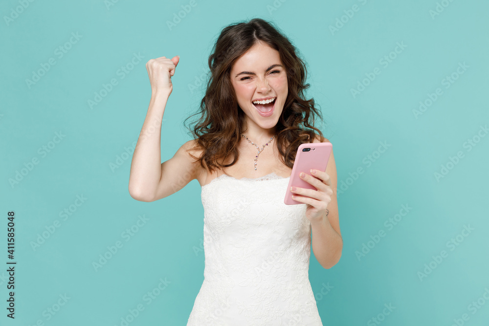 Joyful bride young woman 20s in white wedding dress using mobile cell phone typing sms message doing winner gesture isolated on blue turquoise background studio. Ceremony celebration party concept. Joyful bride young woman 20s in white wedding dress using mobile cell phone typing sms message doing winner gesture isolated on blue turquoise background studio. Ceremony celebration party concept.