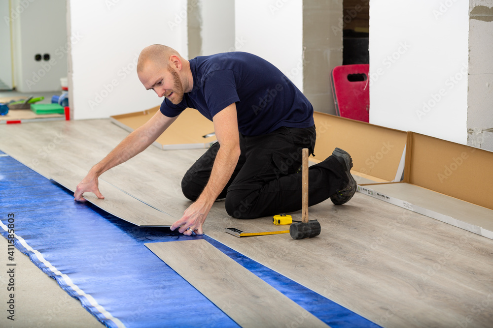 Worker joining click lock vinyl floor plank covering at home renovation. Man laying PVC-floor. Craftsman installing new laminated wooden floor on blue acoustic thermal flooring underlayment. Worker joining click lock vinyl floor plank covering at home renovation. Man laying PVC-floor. Craftsman installing new laminated wooden floor on blue acoustic thermal flooring underlayment.