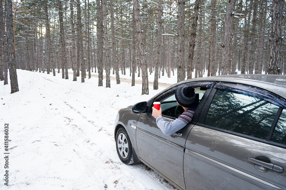 Car on a winter road in the forest. Road trip concept. A young man leans out of the car window and looks at the snowy forest around.