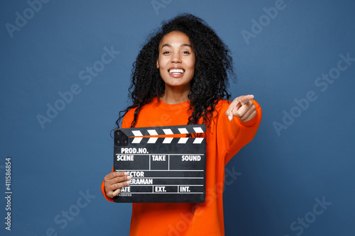Φωτογραφία Cheerful young african american woman in bright orange sweatshirt standing hold classic black film making clapperboard pointing index finger camera on you isolated on blue background studio portrait