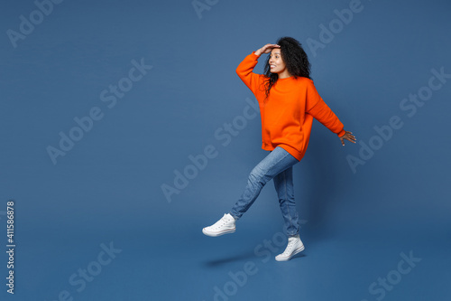 Full length side view of cheerful young african american woman in casual bright orange sweatshirt holding hand at forehead looking far away distance isolated on blue color background studio portrait.