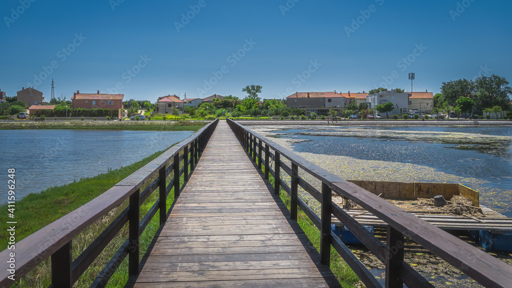 Obraz premium Wooden bridge between fishing ponds or lakes leading to residential suburbs of Nin with clear blue sky on sunny day, Croatia