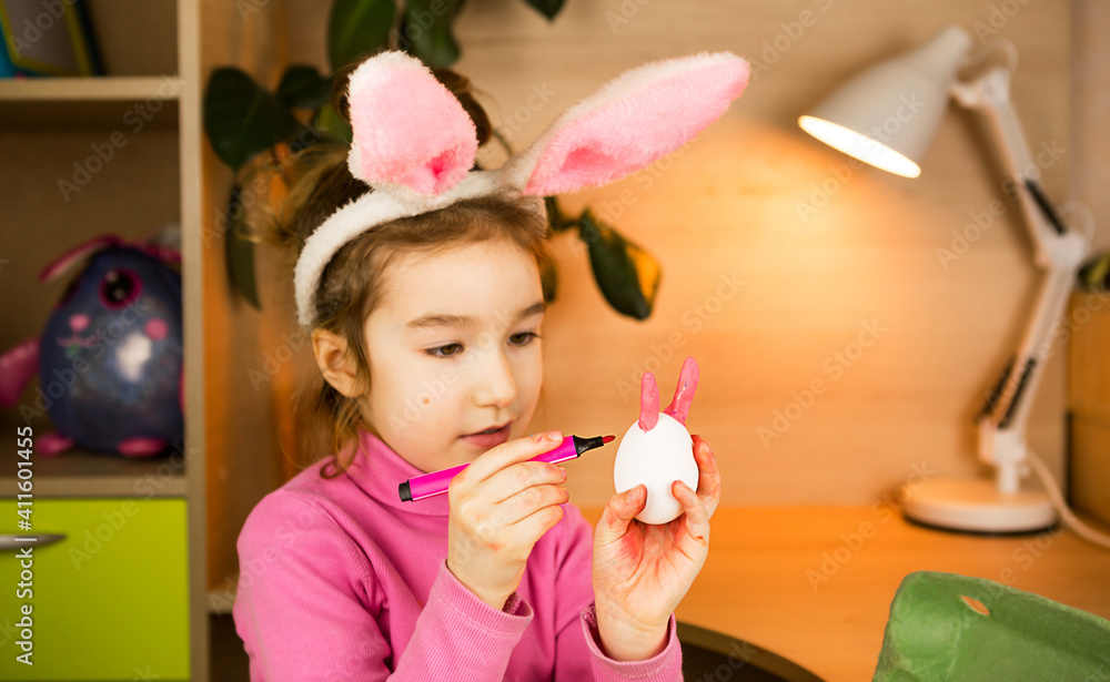 A girl in the ears of an Easter bunny paints eggs with a felt-tip pen in the home interior. Crafts, preparation for a religious holiday, a tray with eggs, hare ears made of plasticine A girl in the ears of an Easter bunny paints eggs with a felt-tip pen in the home interior. Crafts, preparation for a religious holiday, a tray with eggs, hare ears made of plasticine