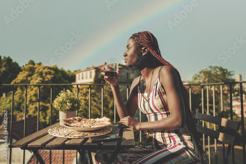 A pensive young dazzling African female in a striped summer jumpsuit is sitting on a balcony of an outdoor restaurant after the rain, drinking wine and eating pizza; a rainbow behind her on the sky