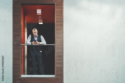 A serious mature businessman in a plaid vest is typing a message via the cellphone while leaning against the chrome railing of a balcony indoors of a modern office with a copy space place on the right © skyNext