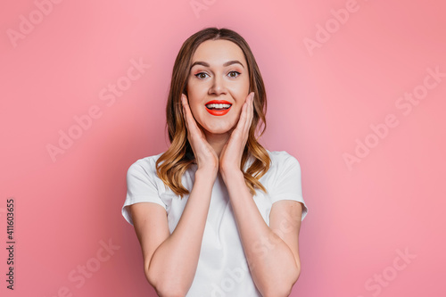 Young caucasian shocked, surprised woman with red lips in a white t-shirt smiling looking at the camera isolated on pink background