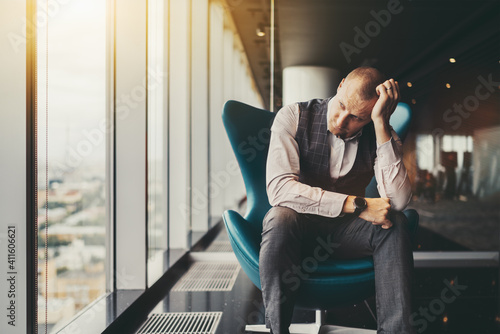 A tired depressed businessman in a luxury office settings sadly looking outside the window; a man entrepreneur on an armchair is worrying about his dismissal and the bankruptcy of the company photo