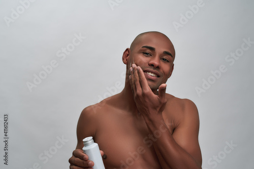 Shirtless handsome african american guy smiling at camera, applying aftershave lotion while posing isolated over gray background photo