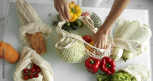 Woman takes yellow and red pepper out from reusable grocery bag with vegetables on a table at the kitchen at home after grocery shopping. Zero waste and plastic free concept. Mesh cotton shopper.