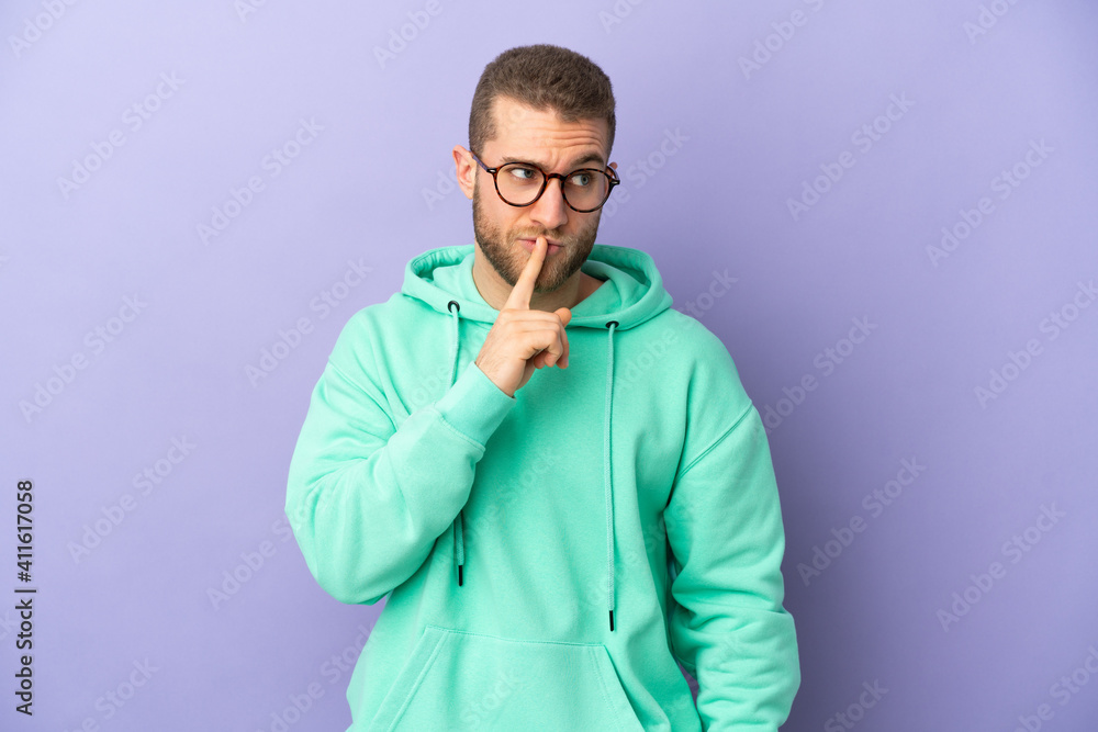 Young handsome caucasian man isolated on purple background showing a sign of silence gesture putting finger in mouth