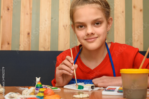 A girl paints a craft from salt dough with watercolors and looked into the frame photo