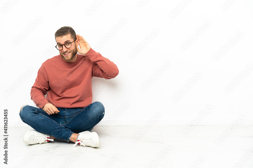 Young handsome man sitting on the floor listening to something by putting hand on the ear