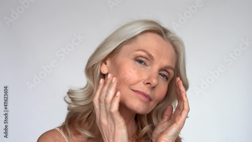 Happy 50s middle aged mature woman touching facial skin looking at camera pampering in mirror. Old healthy dry skin care beauty concept, skincare treatment, cosmetics and anti age plastic surgery.