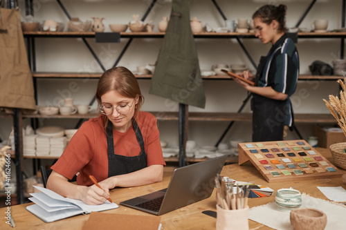 Portrait of female small business owner taking notes in planner while working at wooden table with laptop in pottery workshop, copy space