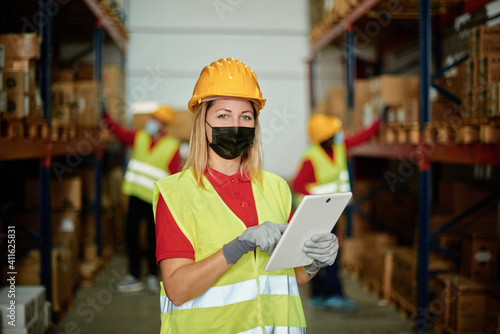 Portrait of happy female worker looking at camera inside warehouse while wearing face mask - Focus on face