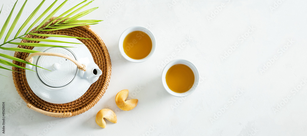 View from above of Asian tea concept, two white cups of tea, fortune cookies and teapot surrounded with dry green tea on light background.Wide banner. - obrazy, fototapety, plakaty 