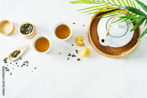View from above of Asian tea concept, two white cups of tea, fortune cookies and teapot surrounded with dry green tea on light background.