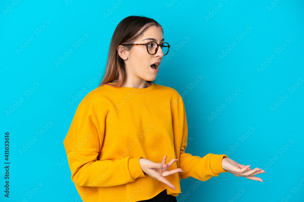 Young caucasian woman isolated on blue background with surprise expression while looking side - obrazy, fototapety, plakaty 