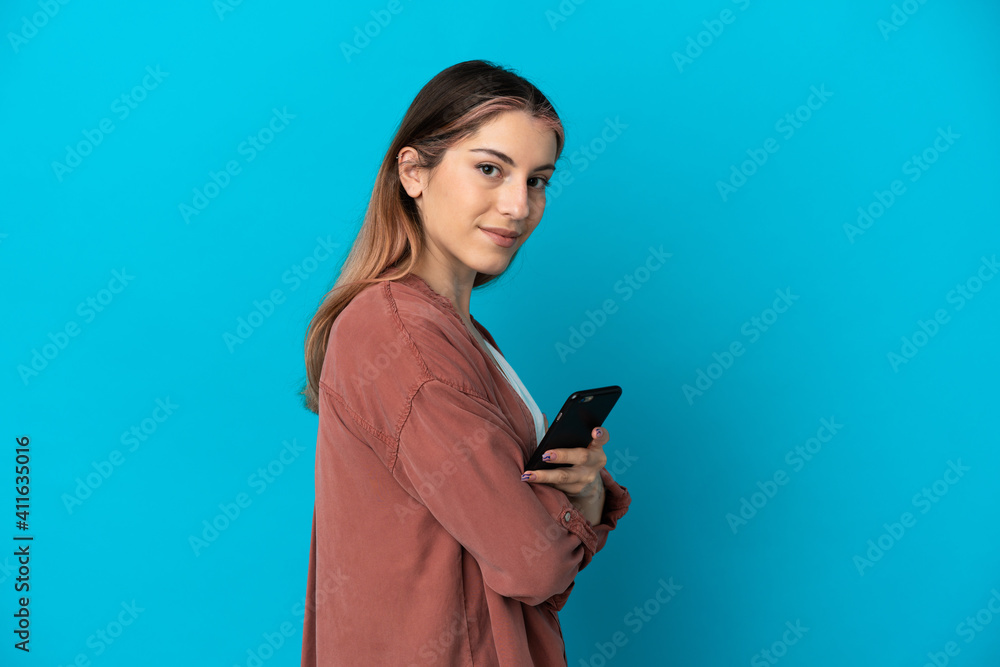 Young caucasian woman isolated on blue background holding a mobile phone and with arms crossed