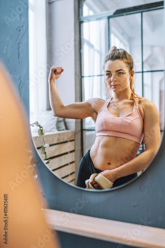 Beautiful fit woman with perfect abdominal muscles and biceps in front of mirror look at her reflection. Liposuction, weight loss, bodybuilding concept