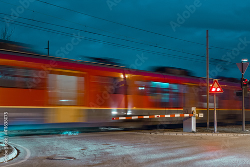Fast moving red train passing grade crossing with barriers down and red light flashing. No cars are visible, only blur of a train over the crossing,