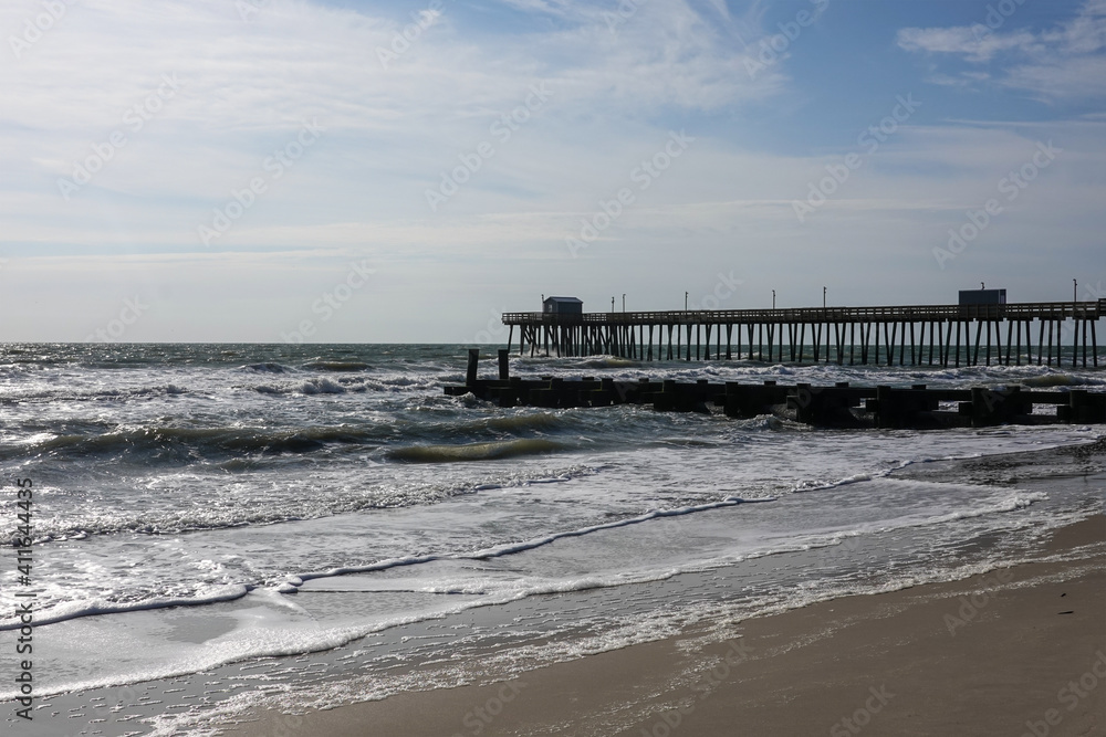 Coastal landscape showing the sandy beach, rough ocean and a pier and wooden jetty extending into the ocean
