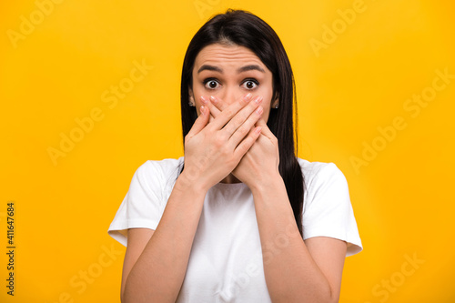 Photo of shocked surprised caucasian brunette girl in basic white t-shirt, standing on isolated orange background, covering her mouth with her hands, looking straight into the camera