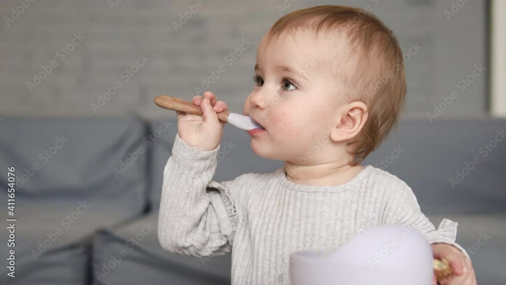 Pretty hungry infant girl eating porridge with spoon from bowl on her own Pretty hungry infant girl eating porridge with spoon from bowl on her own