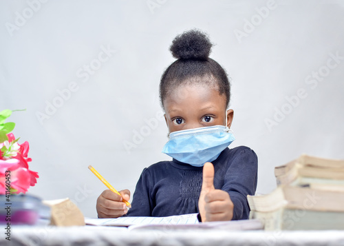 African girl child with nose mask, giving thumbs up while studying alone with books and flowers on her table 