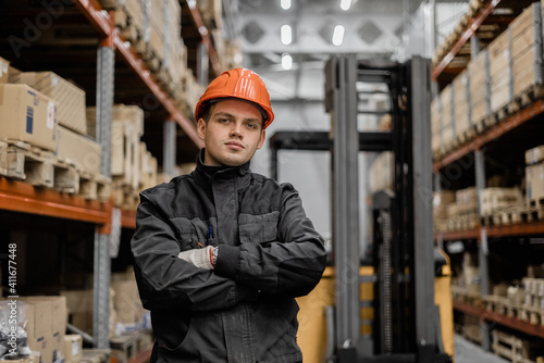 A worker in the background of a warehouse and forklift looks intently into the camera. © CjVitoS