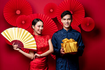 Smiling Asian couple in traditional costumes with fan and gold package as a gift for Chinese new year in oriental style red background