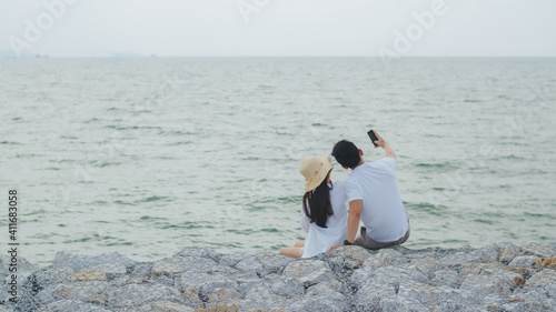 Happy Asian couple using a smartphone take a photo and selfie while sitting on the stone at the seaside during beautiful morning sunrise. Romantic couple travel on vacation weekend.