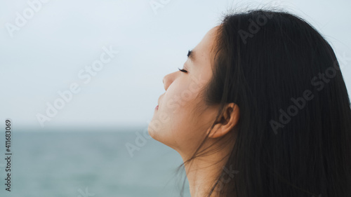Beautiful Asian female traveler keeping eyes closed and breathing fresh air at the ocean view. Health wellness lifestyle concept.