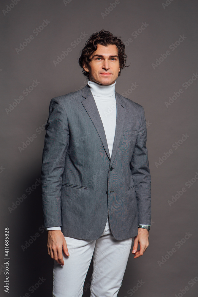 Fototapeta premium Tall handsome man dressed in white turtleneck, white jeans and grey jacket posing on the grey background